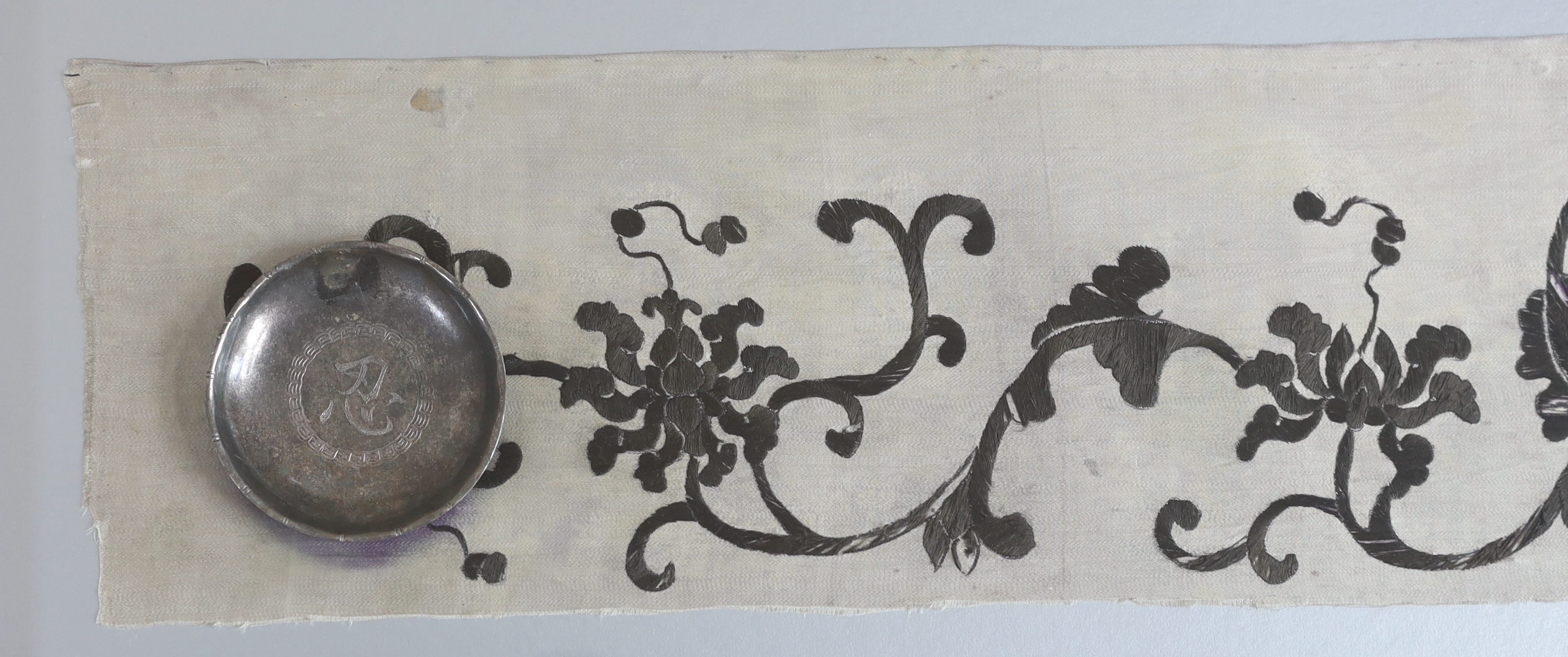 A late 19th century Chinese black and cream silk embroidery, mounted together with an inscribed calligraphic stone, a pair of white metal chop sticks and a similar dish, embroidery: 5.5cm wide, 105cm long